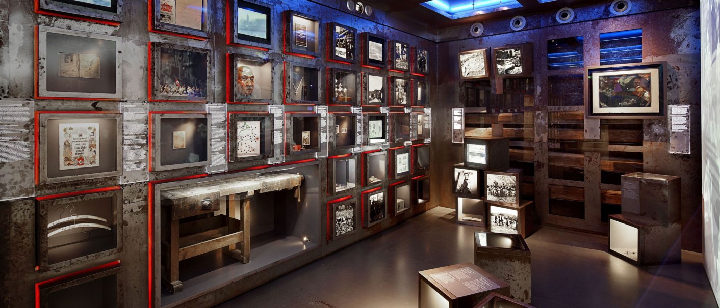 An exhibition room with illuminated showcases and pictures in the House of History Baden-Württemberg, themed around the world wars., © danielstauch_gf