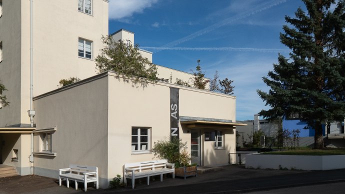 Beige Architekturgalerie mit Flachdach, B&auml;nken und gro&szlig;em Nadelbaum vor blauem Himmel., &copy; Thomas F&uuml;tterer