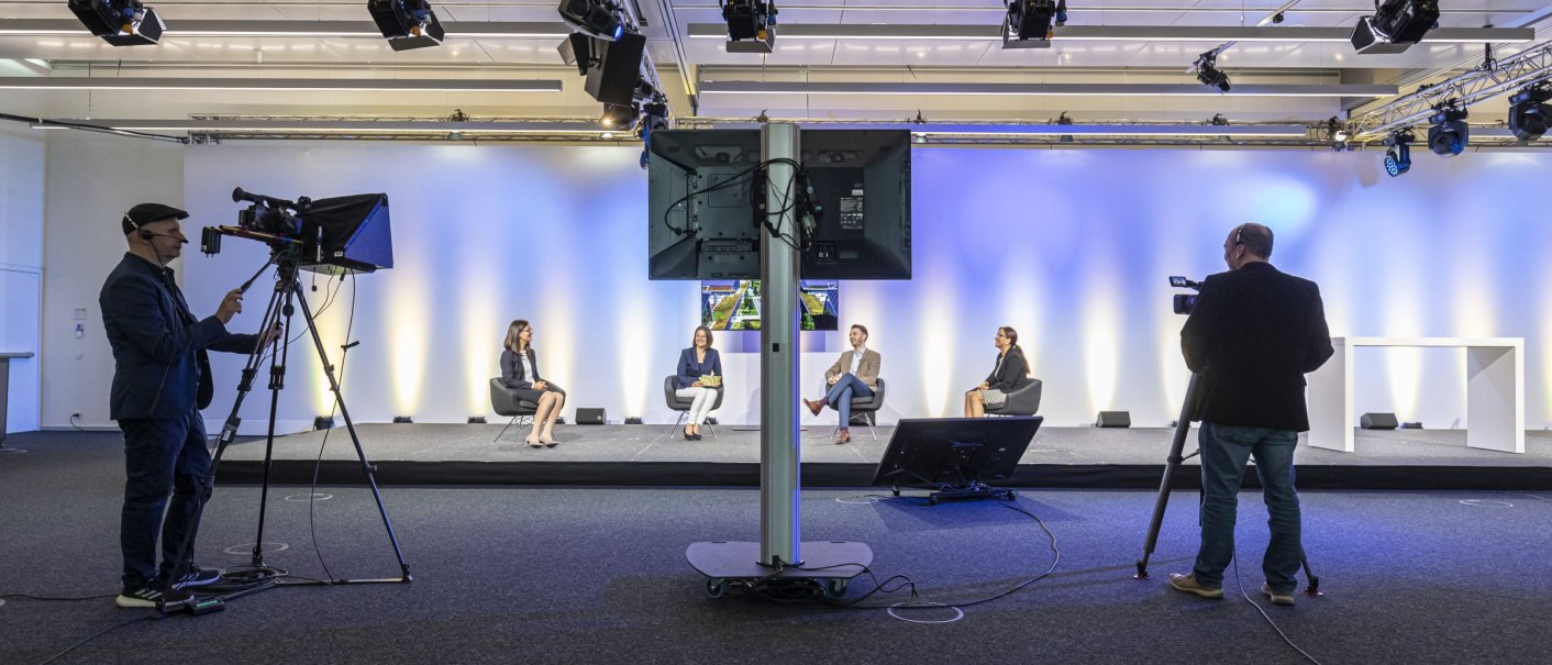 Ein modernes Streaming-Studio mit Kameraleuten und Personen auf einer Bühne. Die Wand ist blau-gelb beleuchtet, und es gibt mehrere Kameras und Monitore., © Messe Stuttgart