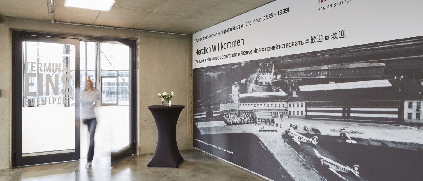 Eingangsbereich der Motorworld Region Stuttgart mit einem großen historischen Foto an der Wand und einer Glastür mit der Aufschrift 'Terminal Eins'., © MW Region Stuttgart