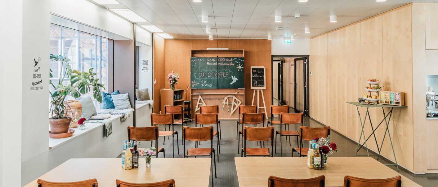 Moderner Raum mit Stühlen, Tischen und einer Tafel. Pflanzen und Kissen auf der Fensterbank, Snacks auf einem Tisch. Schriftzug "Out of Office" auf der Tafel., © OutOfOffice GmbH Moderner Raum mit Stühlen, Tischen und einer Tafel. Pflanzen und Kissen auf der Fensterbank, Snacks auf einem Tisch. Schriftzug "Out of Office" auf der Tafel., © OutOfOffice GmbH