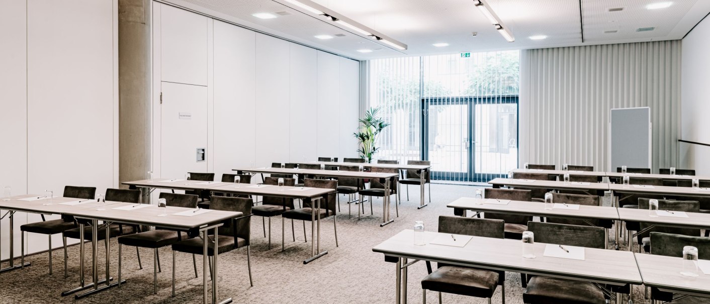 Moderner Tagungsraum mit Tischen und Stühlen, einem Deckenprojektor und großen Fenstern mit Jalousien. Helle, neutrale Farben dominieren den Raum., © VHE Mo.Stuttgart