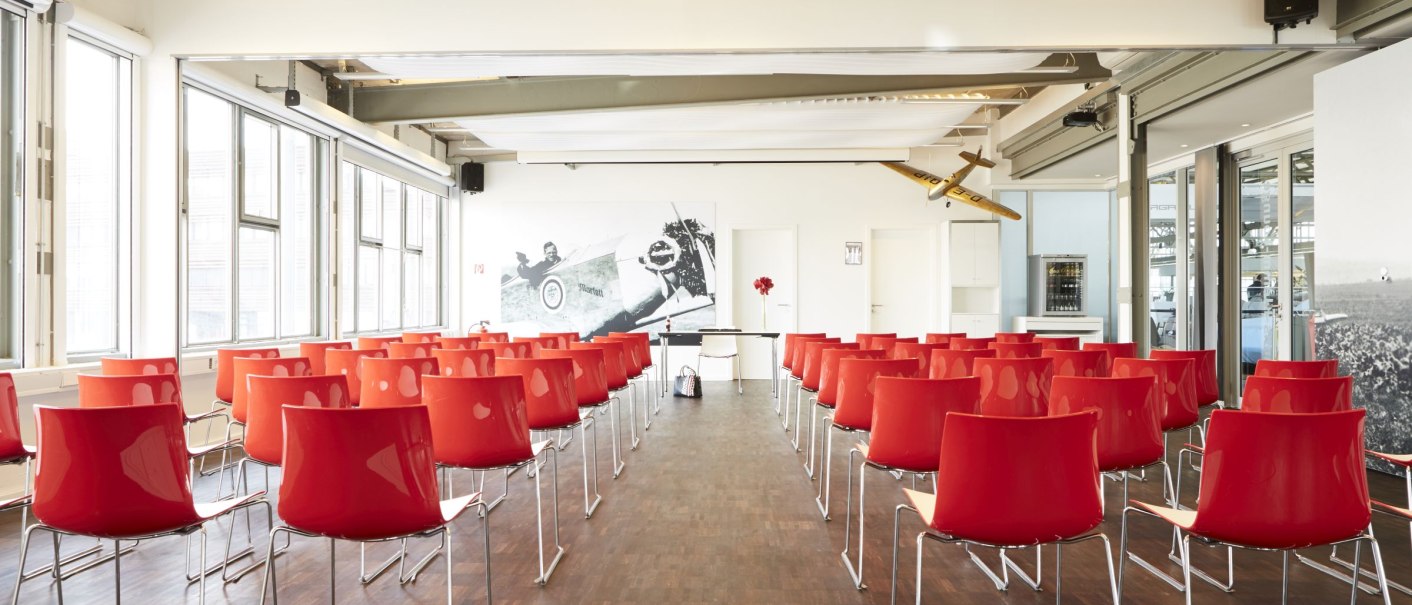 Ein heller Konferenzraum mit roten Stühlen, Holzboden und einem großen Rennwagenbild an der Wand. An der Decke hängt ein Modellflugzeug., © MW Region Stuttgart