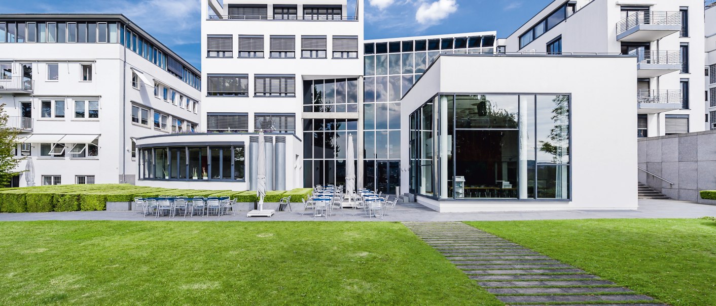 Modernes Gebäude mit Glasfassade und Rasenfläche, umgeben von weiteren weißen Gebäuden. Klare Linien und große Fenster prägen die Architektur., © Francis Koenig Modernes Gebäude mit Glasfassade und Rasenfläche, umgeben von weiteren weißen Gebäuden. Klare Linien und große Fenster prägen die Architektur., © Francis Koenig