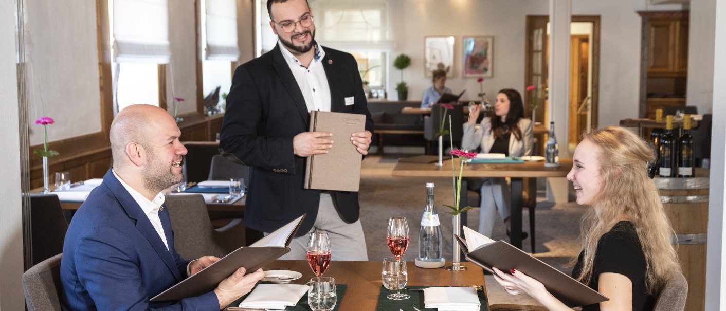 A couple is sitting in an elegant restaurant, smiling with menus. A waiter stands next to them. Other guests can be seen in the background., &copy; Schlosshotel Monrepos