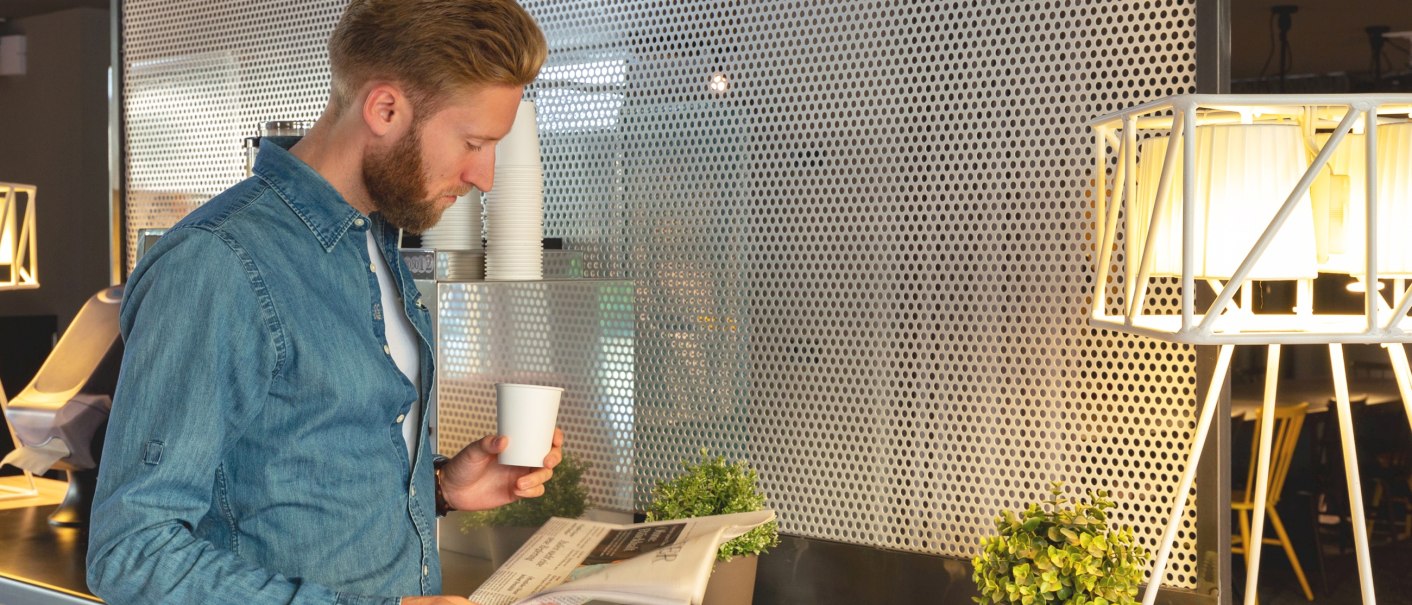 Ein Mann in einem Jeanshemd liest eine Zeitung in einer modernen Lobby. Er hält einen Kaffeebecher, umgeben von Pflanzen und einer Lampe., © a&o Hostels Ein Mann in einem Jeanshemd liest eine Zeitung in einer modernen Lobby. Er hält einen Kaffeebecher, umgeben von Pflanzen und einer Lampe., © a&o Hostels