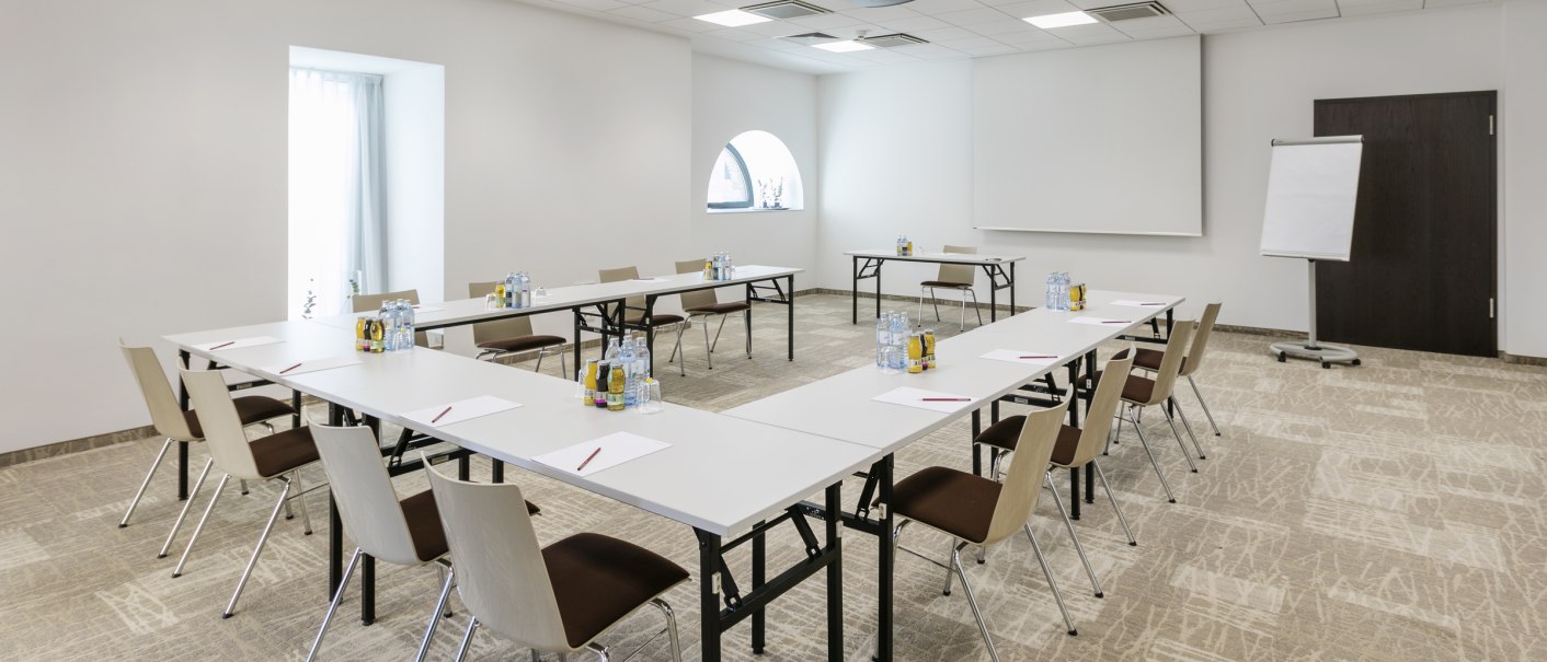 Moderner Tagungsraum mit U-förmiger Tischanordnung, Stühlen, Flipchart und Projektor. Helle Wände und Teppichboden schaffen eine professionelle Atmosphäre., © ARCOTEL Hotels