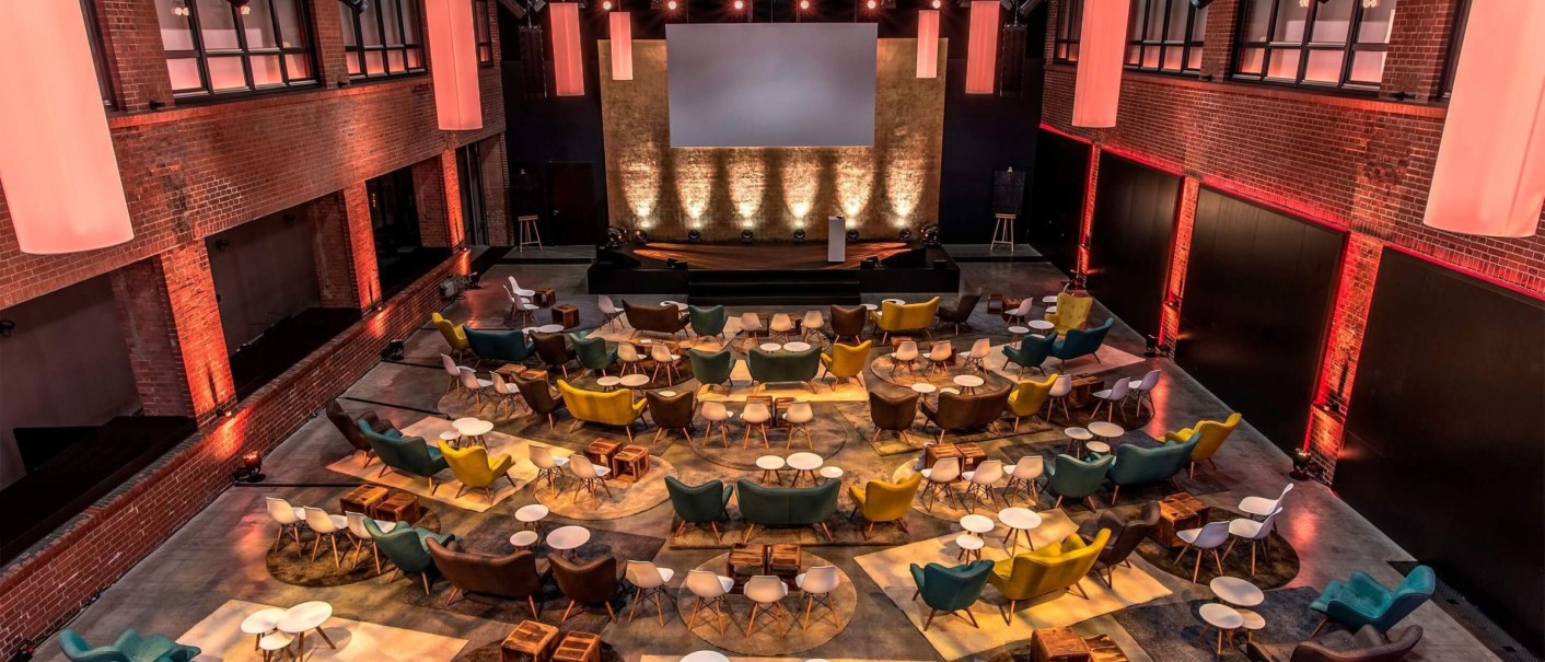 Moderner Veranstaltungsraum mit bunten Sesseln und Tischen, Bühne und Leinwand. Ziegelwände und warme Beleuchtung schaffen eine einladende Atmosphäre., © rauschenbergercatering