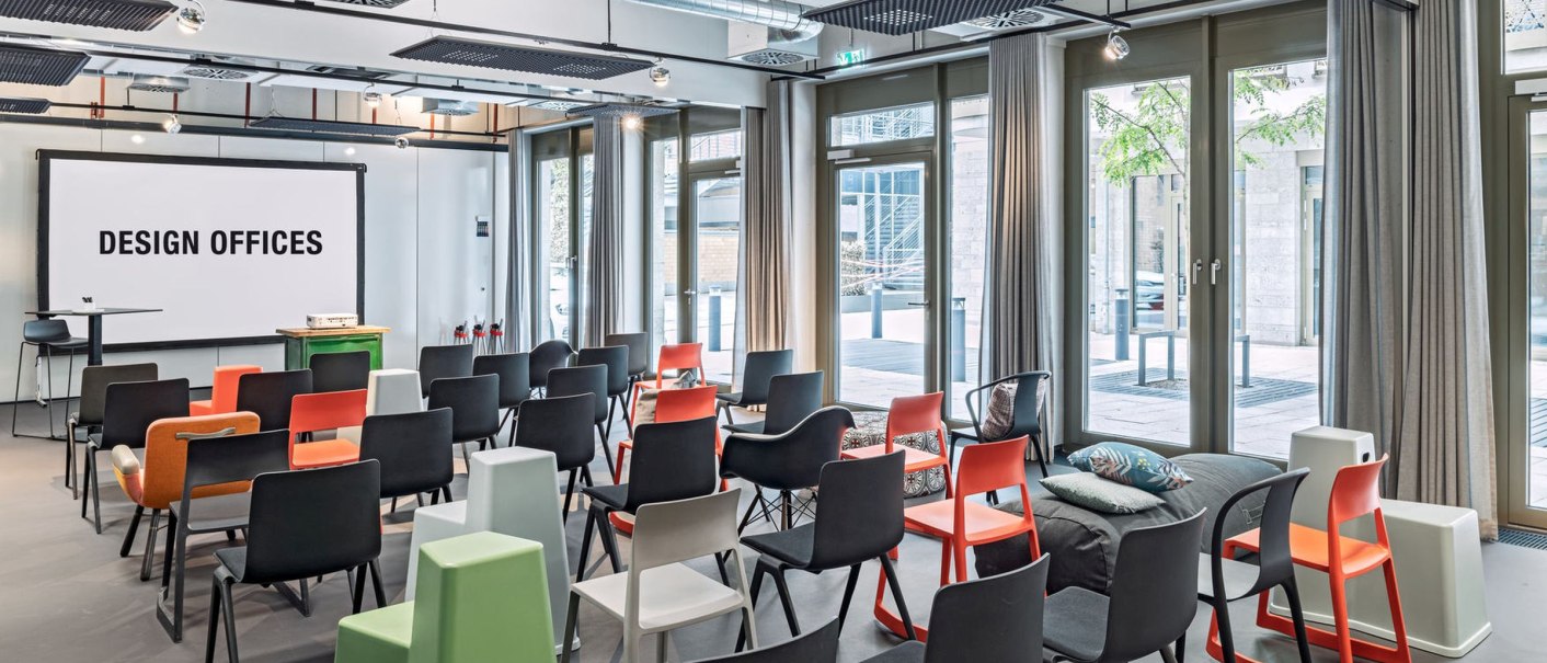 Moderner Seminarraum mit bunten Stühlen, Leinwand und großen Fenstern. Industrielles Design mit sichtbaren Lüftungsrohren und moderner Einrichtung., © Design Offices