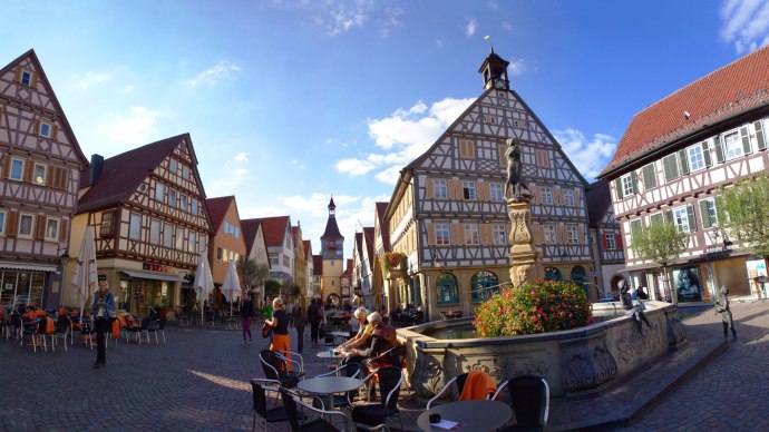 Half-timbered houses in Winnenden with a fountain in the foreground. People sit and walk on the cobbled square under a blue sky., &copy; SMG_Achim Mende