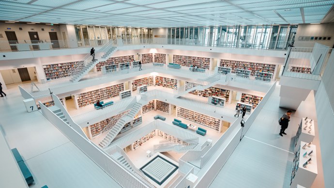 Moderne, helle Bibliothek mit mehreren Etagen, weißen Wänden und Bücherregalen. Menschen bewegen sich auf den Treppen und lesen Bücher., © Stuttgart-Marketing GmbH, Sarah Schmid