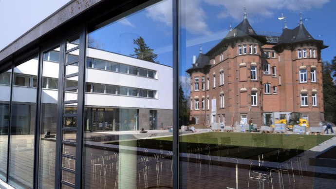 Modern glass fa&ccedil;ade of the Evangelical Academy Bad Boll reflects a historic building and the blue sky. Chairs can be seen in the foreground., &copy; Eabb_Giacinto_Carlucci