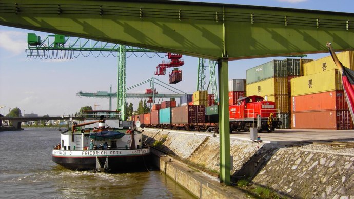 Ein Frachtschiff im Hafen Stuttgart neben Containern und einem Güterzug. Kräne und Wasser im Hintergrund, klare Himmel., © Hafen Stuttgart GmbH