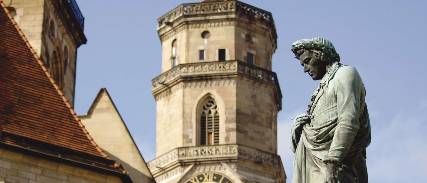 Das Schillerdenkmal steht vor einem historischen Turm mit Uhr und gotischen Fenstern, unter einem klaren blauen Himmel., © Stuttgart-Marketing GmbH Das Schillerdenkmal steht vor einem historischen Turm mit Uhr und gotischen Fenstern, unter einem klaren blauen Himmel., © Stuttgart-Marketing GmbH
