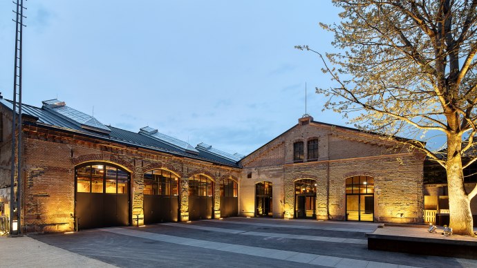 Beleuchtete Wagenhallen mit Backsteinfassade bei D&auml;mmerung, Baum im Vordergrund. Historische Architektur mit modernen Elementen., &copy; Wagenhallen Stuttgart GmbH & Co. KG