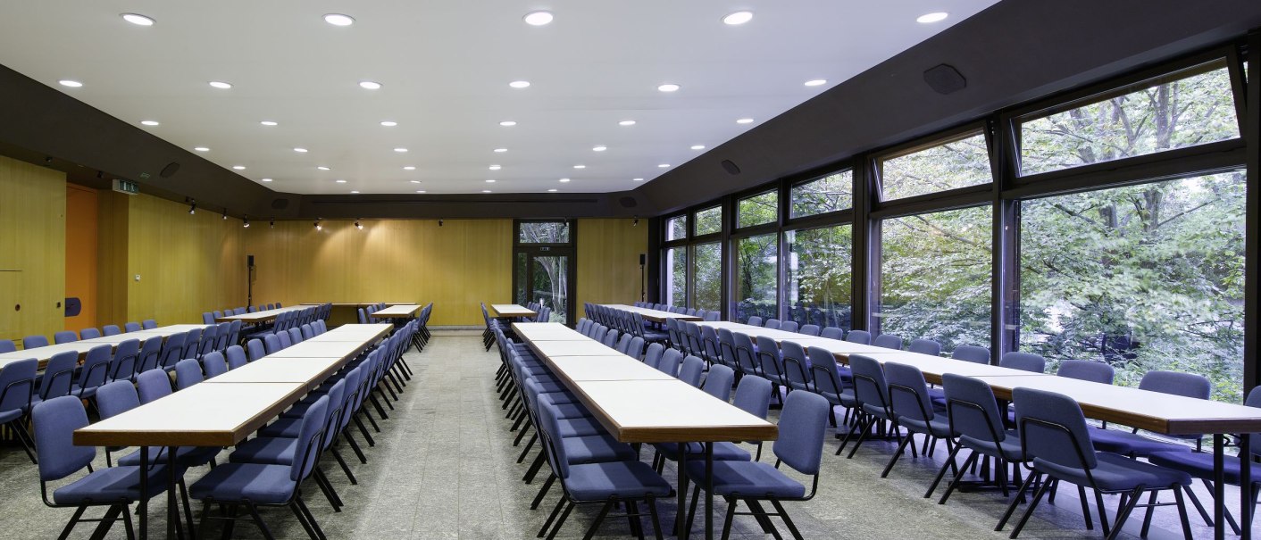 Ein heller Konferenzraum mit langen Tischen und blauen Stühlen, großen Fenstern und moderner Beleuchtung., © eslive Ein heller Konferenzraum mit langen Tischen und blauen Stühlen, großen Fenstern und moderner Beleuchtung., © eslive