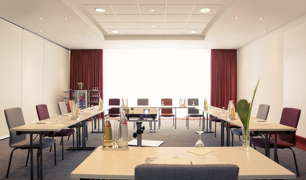 A bright conference room with a U-shaped table arrangement, chairs, drinks and a projector. Red curtains and white walls create a professional atmosphere., &copy; Mercure Stuttgart Airport Messe