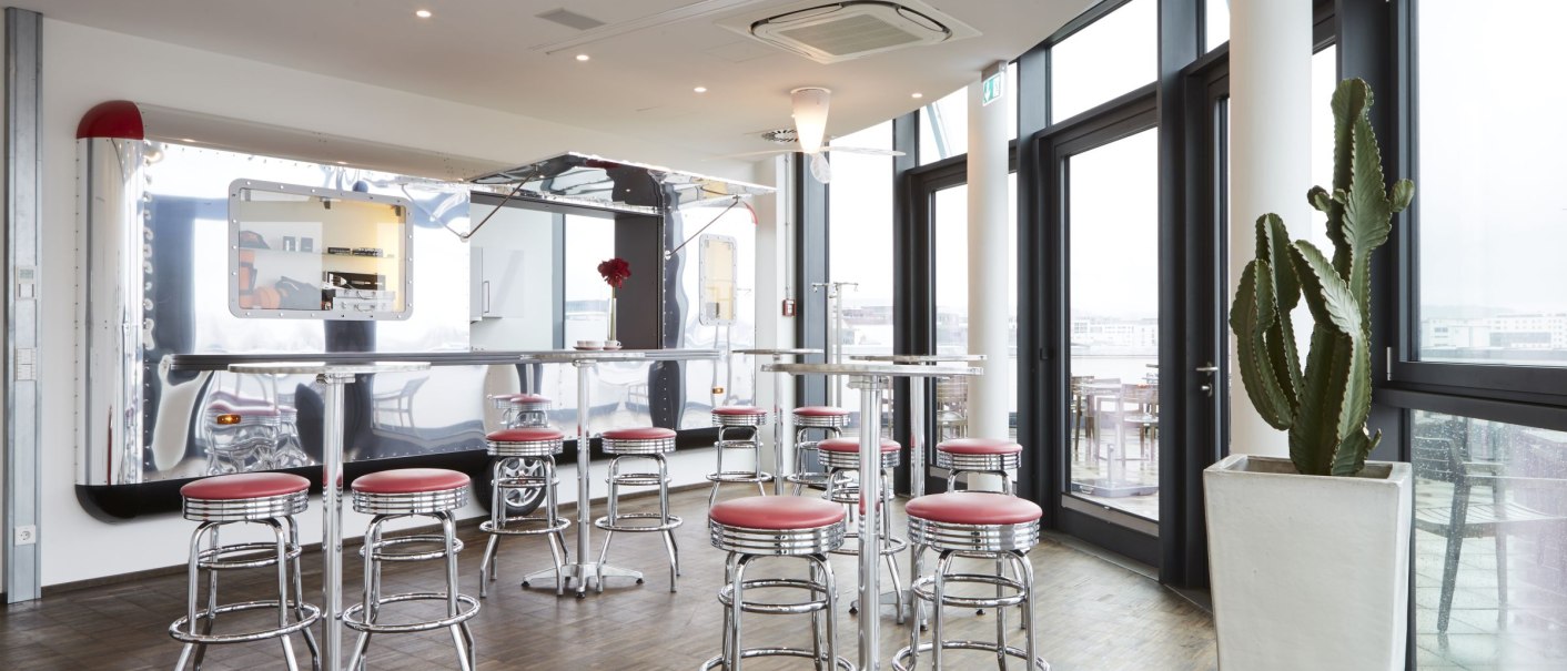 Modernes Café mit Airstream-Design, roten Barhockern und großen Fenstern. Ein Kaktus steht in einem weißen Topf., © MW Region Stuttgart