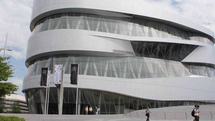 Das Mercedes-Benz Museum in Stuttgart zeigt seine moderne, geschwungene Architektur mit Glasfassade und mehreren Flaggen davor., © Stuttgart-Marketing GmbH