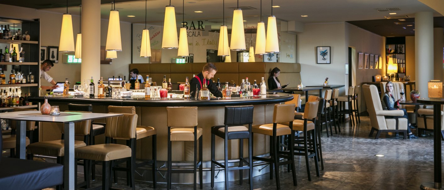 Stilvolle Hotelbar mit runder Theke, Barhockern und moderner Beleuchtung. G&auml;ste sitzen entspannt, w&auml;hrend ein Barkeeper arbeitet., &copy; ARCOTEL Hotels