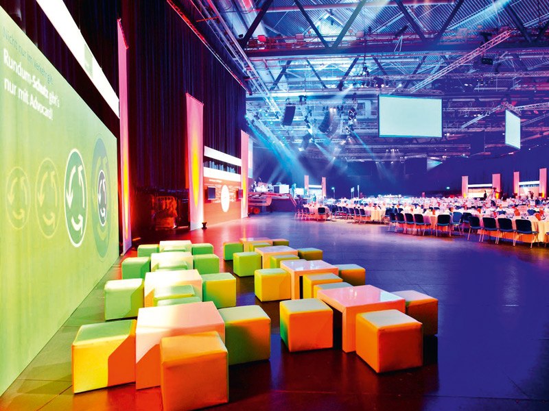 Bunte Sitzwürfel und gedeckte Tische in der Hanns-Martin-Schleyer-Halle, beleuchtet in Blau und Orange., © Timo Deiner