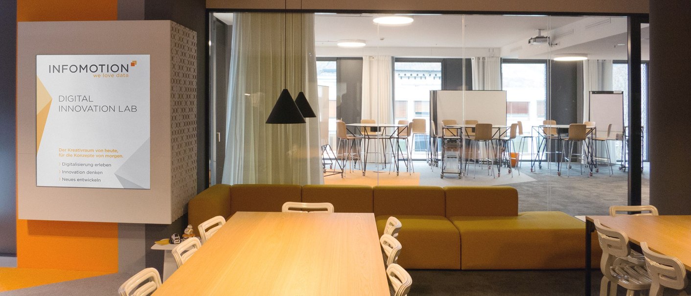 Modern room with wooden tables, white chairs and yellow sofas. A sign reads 'INFOMOTION Digital Innovation Lab'. A meeting room in the background., © INFOMOTION
