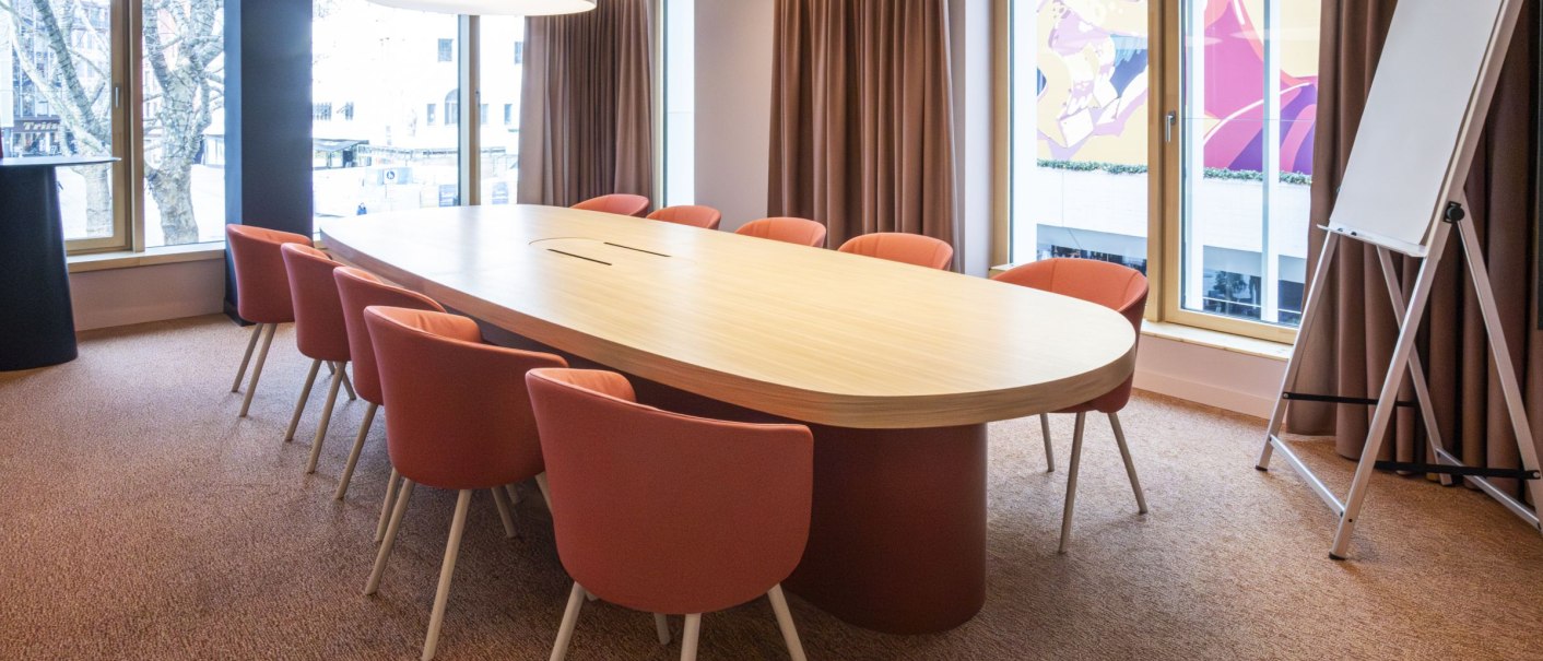 A modern conference room with a large oval wooden table surrounded by red chairs. Large windows provide plenty of daylight. A flipchart is available., &copy; SMG Stuttgart Marketing GmbH - Sarah Schmid