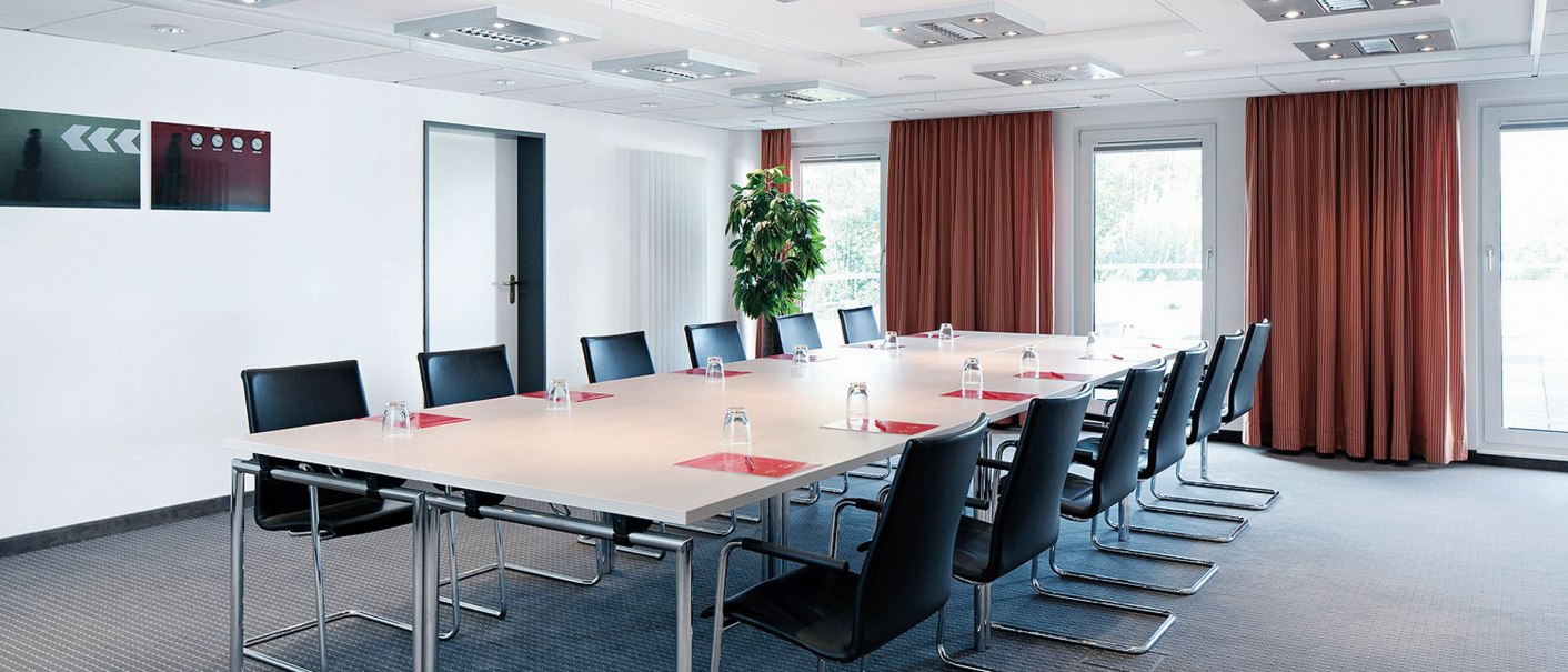 Moderner Konferenzraum mit großem Tisch, schwarzen Stühlen und Gläsern. Helle Beleuchtung, rote Vorhänge und eine Pflanze im Hintergrund., © Seminaris Hotels