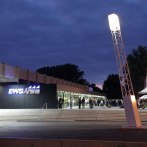 The EWS Arena at dusk, people standing in front of the entrance. An illuminated pole and a bicycle stand are visible., © Dietmar Strauss