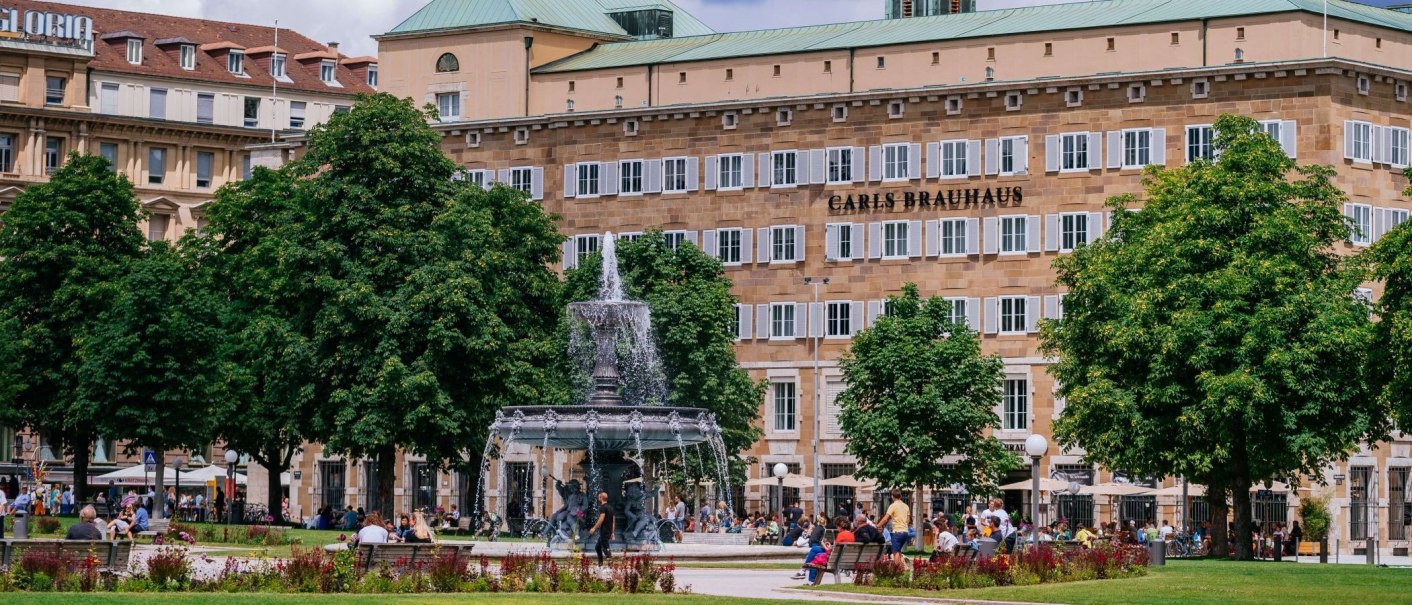 Belebter Schlossplatz mit Brunnen, umgeben von Bäumen und Menschen. Im Hintergrund ein Gebäude mit 'Carls Brauhaus'., © Thomas Niedermüller