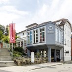 The Backnang community center with a modern façade, large windows and two colorful flags. A staircase leads to the entrance., © © die arge lola, Kai Loges + Andreas Langen