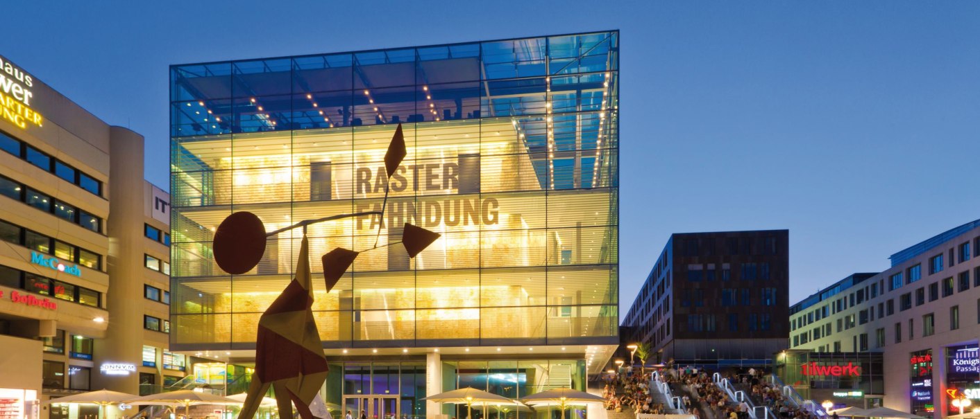 Das Kunstmuseum Stuttgart bei Abenddämmerung, beleuchtet mit moderner Skulptur im Vordergrund und Menschen auf der Treppe daneben., © SMG, Werner Dieterich