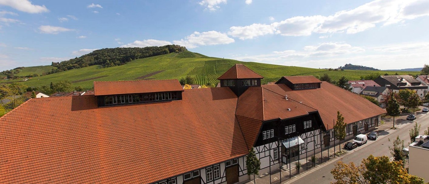 Die Alte Kelter in Fellbach mit rotem Ziegeldach und Fachwerk, umgeben von grünen Weinbergen und blauem Himmel., © Achim Mende Die Alte Kelter in Fellbach mit rotem Ziegeldach und Fachwerk, umgeben von grünen Weinbergen und blauem Himmel., © Achim Mende