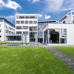 Modernes Gebäude mit Glasfassade und Rasenfläche, umgeben von weiteren weißen Gebäuden. Klare Linien und große Fenster prägen die Architektur., © Francis Koenig