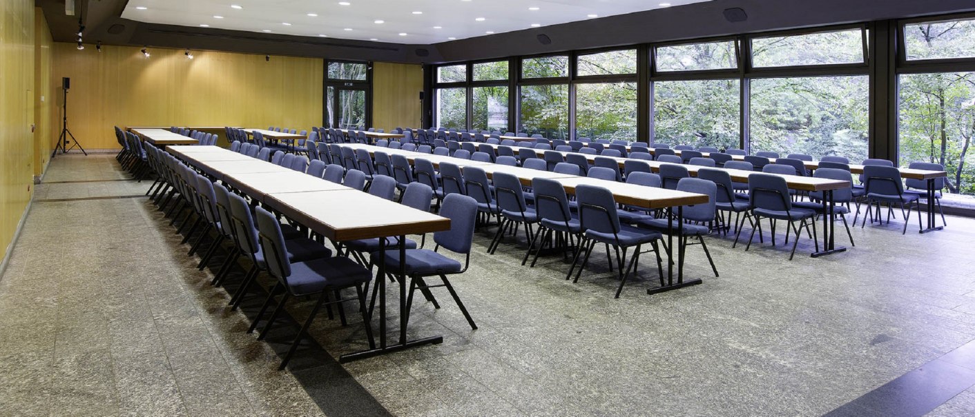 Ein leerer Konferenzraum mit langen Tischen und blauen Stühlen, großen Fenstern und heller Beleuchtung., © eslive Ein leerer Konferenzraum mit langen Tischen und blauen Stühlen, großen Fenstern und heller Beleuchtung., © eslive