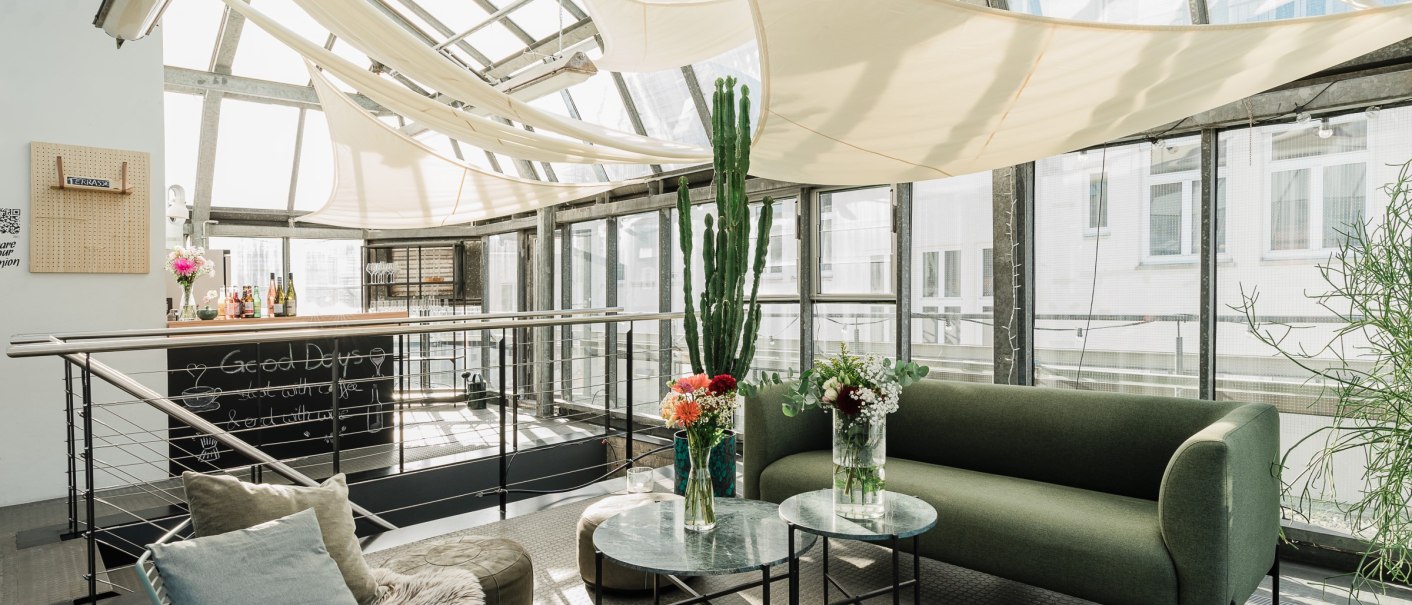 A bright conservatory with a glass roof, green couch, plants and decorative tables. A small bar in the background., © OutOfOffice GmbH