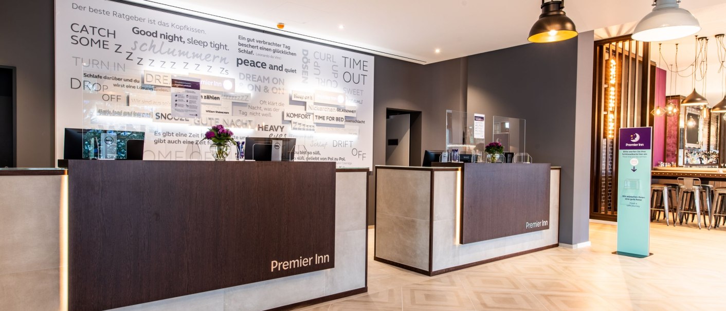 Moderne Hotelrezeption mit zwei Empfangstresen, dekorativer Wand mit Schriftzügen und stilvollen Lampen. Premier Inn Logo sichtbar., © Guenter Lenz Moderne Hotelrezeption mit zwei Empfangstresen, dekorativer Wand mit Schriftzügen und stilvollen Lampen. Premier Inn Logo sichtbar., © Guenter Lenz