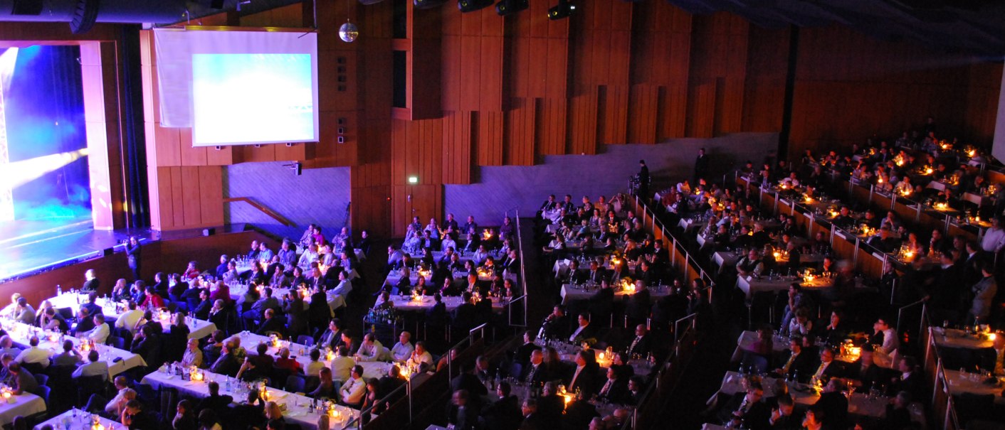 Ein großer Saal mit vielen Menschen, die an Tischen bei Kerzenlicht sitzen. Eine Bühne und eine Leinwand sind sichtbar., © Congress-Centrum Stadtgarten Schwäbisch Gmünd