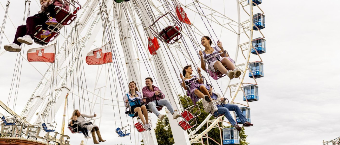 Menschen in Trachten fahren auf einem Kettenkarussell vor einem Riesenrad. Im Hintergrund wehen Fahnen. Volksfestatmosphäre in Stuttgart., © Stuttgart-Marketing GmbH, Sarah Schmid Menschen in Trachten fahren auf einem Kettenkarussell vor einem Riesenrad. Im Hintergrund wehen Fahnen. Volksfestatmosphäre in Stuttgart., © Stuttgart-Marketing GmbH, Sarah Schmid