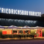 Das Friedrichsbau Varieté bei Nacht, beleuchteter Schriftzug, Sonnenschirme und Tische im Vordergrund., © Friedrichsbau Varieté