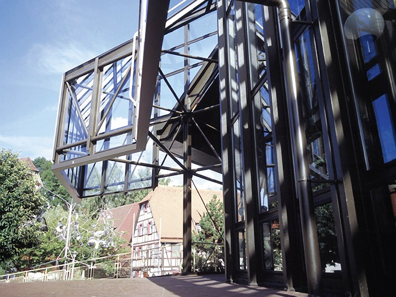 A modern glass facade with geometric patterns, with traditional half-timbered houses and trees in the background under a blue sky., © Stadt Sindelfingen A modern glass facade with geometric patterns, with traditional half-timbered houses and trees in the background under a blue sky., © Stadt Sindelfingen