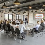 Ein moderner Konferenzraum mit Menschen, die an Tischen sitzen. Ein Bildschirm zeigt das Logo von Erlebnispark Tripsdrill., © Claudia Fy