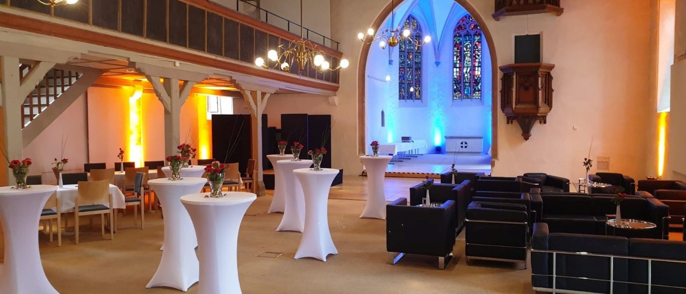 Empfangsbereich der Kreuzkirche Nürtingen mit Stehtischen, Sofas und bunter Beleuchtung. Historische Architektur mit Buntglasfenstern., © kkmaisch_gf