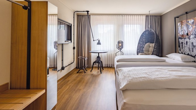Modernes Hotelzimmer mit zwei Betten, H&auml;ngesessel, Tisch und Fernseher. Gro&szlig;e Fenster mit Vorh&auml;ngen lassen viel Licht herein., &copy; Seminaris Hotels (Gerhard Hinz)