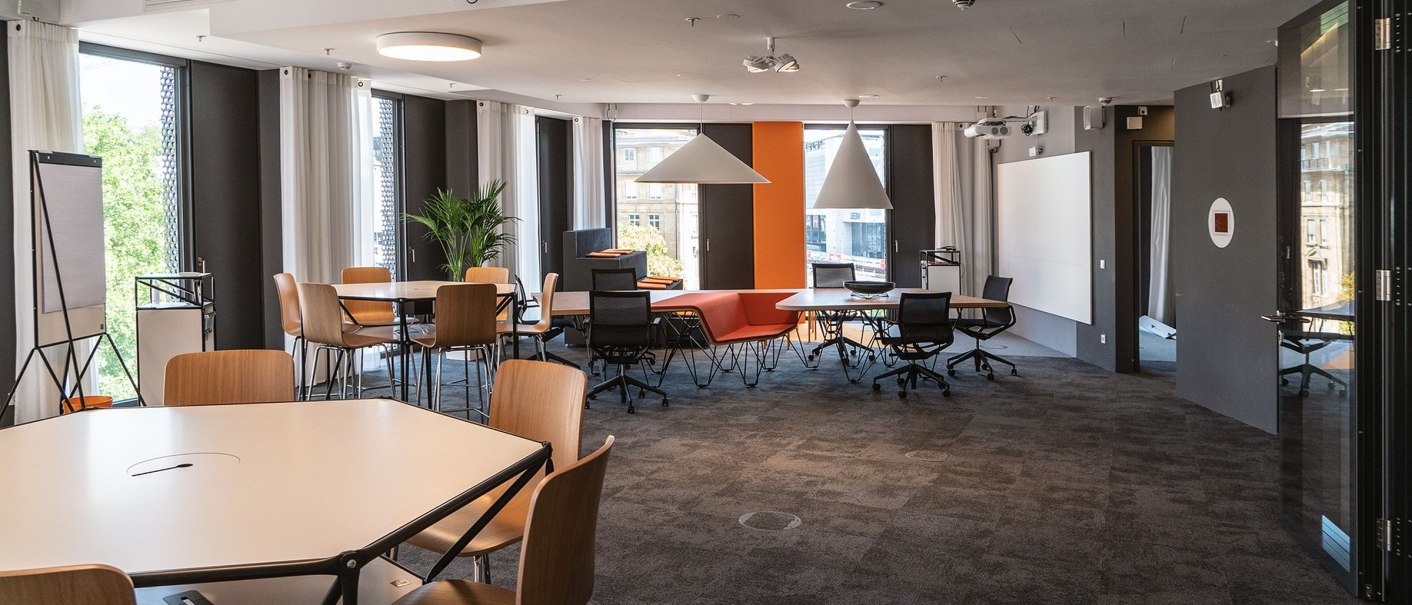 Moderner Arbeitsbereich mit Tischen, Stühlen und großen Fenstern. Helle Beleuchtung, Pflanzen und eine orangefarbene Wand schaffen eine kreative Atmosphäre., © INFOMOTION