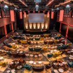 Modern event room with colorful armchairs and tables, stage and screen. Brick walls and warm lighting create an inviting atmosphere., © rauschenbergercatering