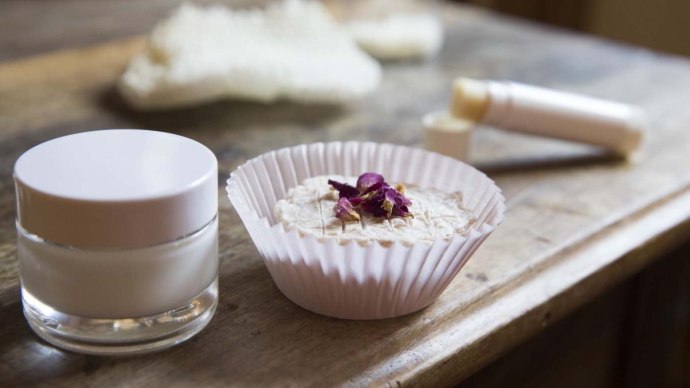 Ein Glas mit Creme, ein festes Kosmetikprodukt in einem Papierförmchen und ein Lippenpflegestift auf einem Holztisch., © hoponopo GmbH