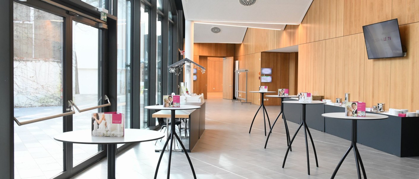 Modernes Foyer mit hohen Fenstern, Holzwänden und Stehtischen. Ein Fernseher hängt an der Wand, und Broschüren liegen auf den Tischen., © Guenter E. Bergmann - Photography