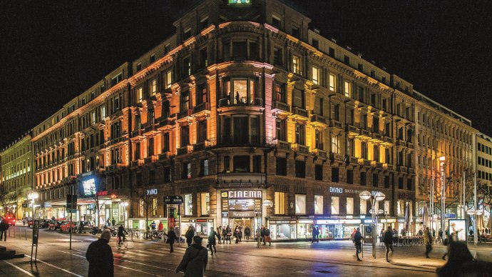 Ein beleuchtetes Geb&auml;ude bei Nacht mit einem Kino und Gesch&auml;ften im Erdgeschoss. Menschen gehen auf der Stra&szlig;e vorbei., &copy; Schauspielb&uuml;hnen Stuttgart