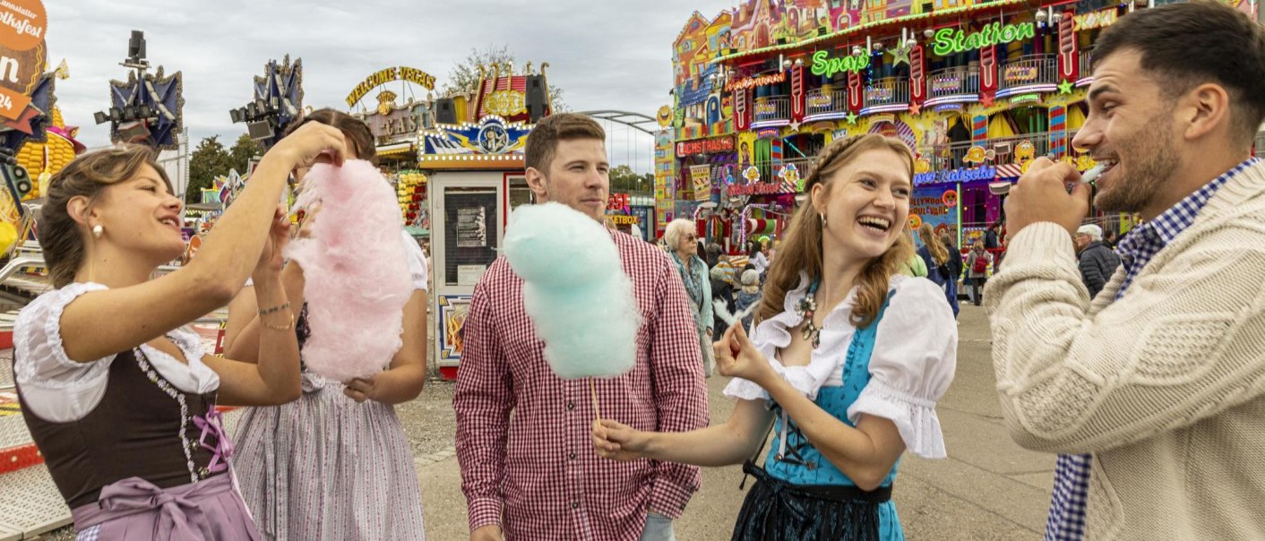 Menschen in Tracht genie&szlig;en Zuckerwatte auf einem bunten Volksfest in Stuttgart. Im Hintergrund sind Fahrgesch&auml;fte und bunte Lichter zu sehen., &copy; Stuttgart-Marketing GmbH, Sarah Schmid